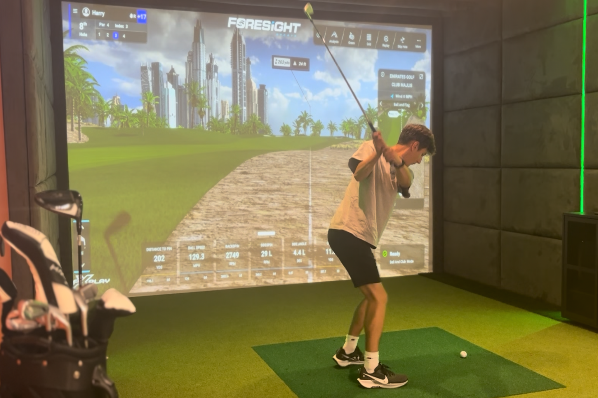 A man is playing golf in an indoor simulator. He is mid-swing. Skyscrapers, palm trees, and a sandy path ARE SHOWN ON A LARGE SCREEN. Golf statistics and game interface details are visible on the screen. To the left, a golf bag with clubs is partially visible.